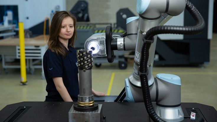 Woman monitoring a Universal Robots collaborative robot arm using a vision system to inspect a mechanical part in an industrial lab.