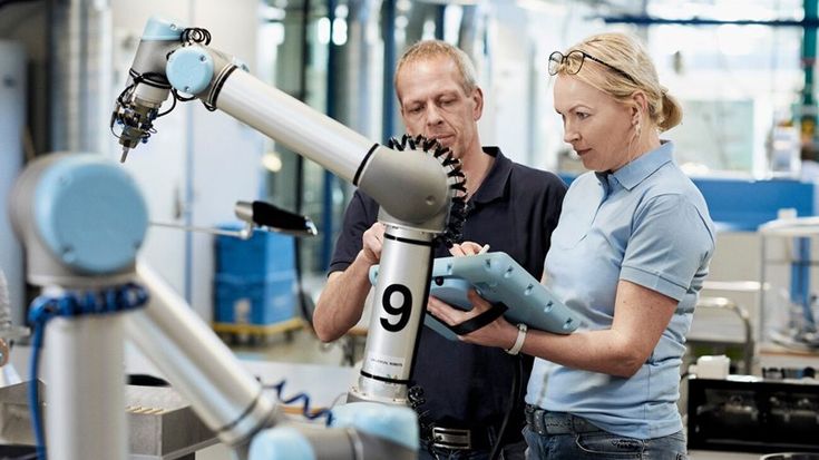 Two technicians program a Universal Robots collaborative robot arm on a factory floor using a teach pendant.