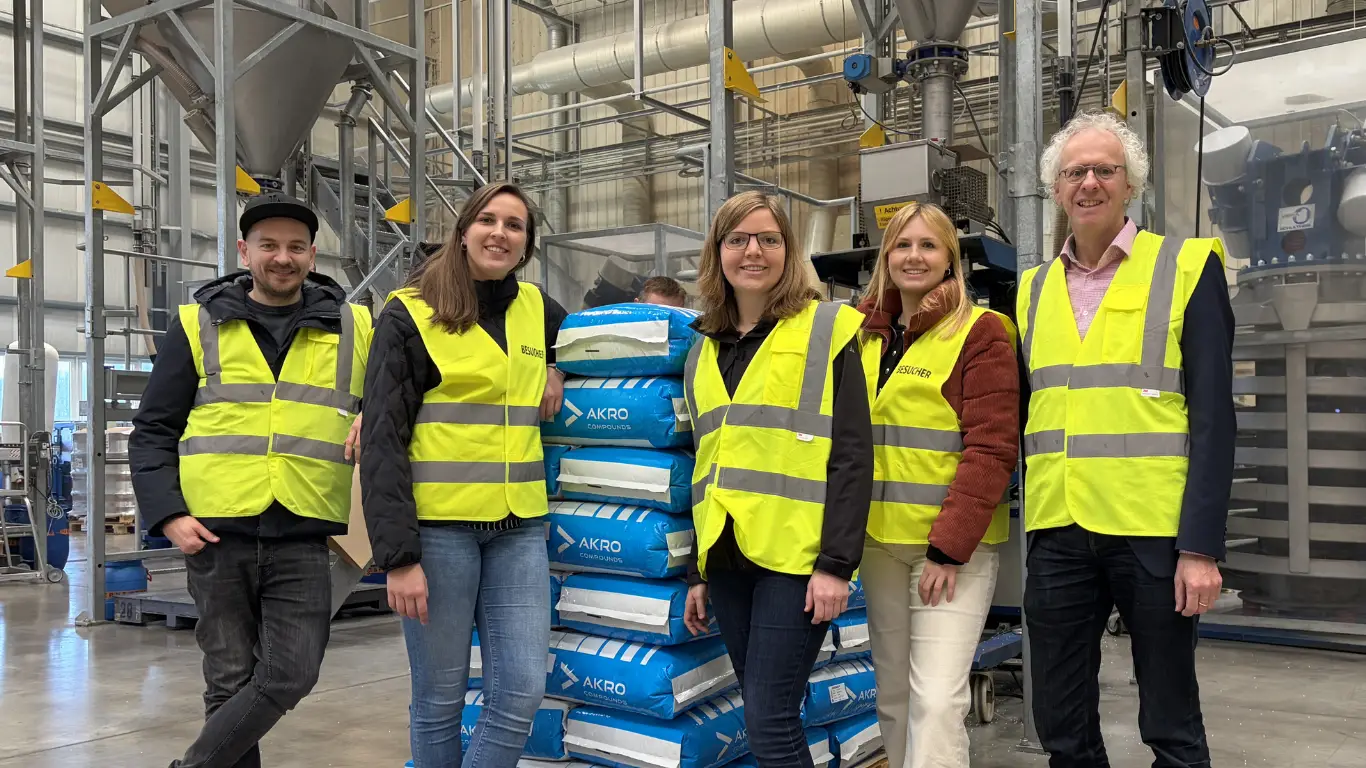 Five people in yellow safety vests stand in an industrial facility, smiling in front of stacked bags labeled "AKRO Compounds."
