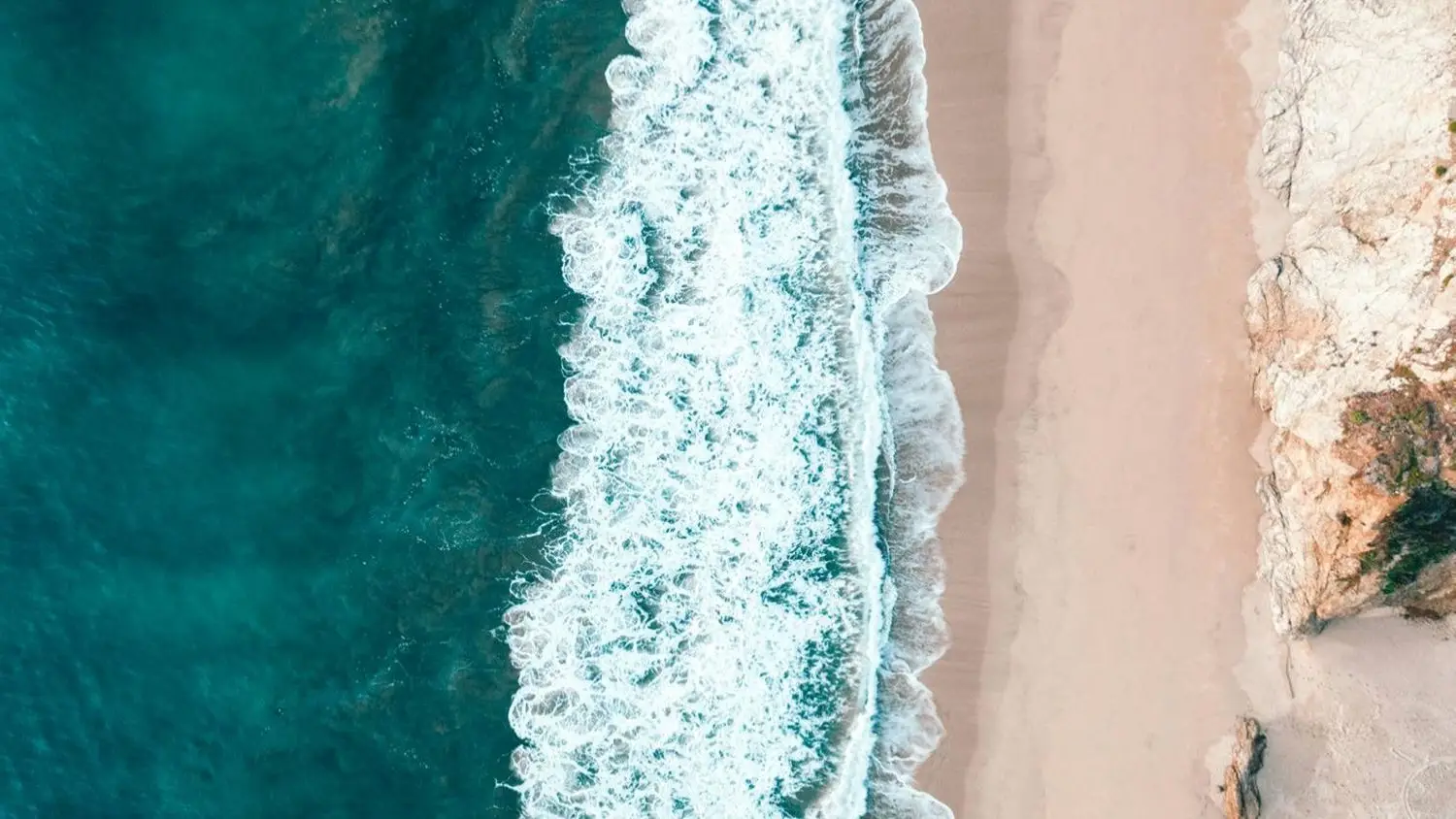 Aerial view of ocean waves crashing on a sandy beach beside rocky cliffs, with vibrant blue and turquoise water.