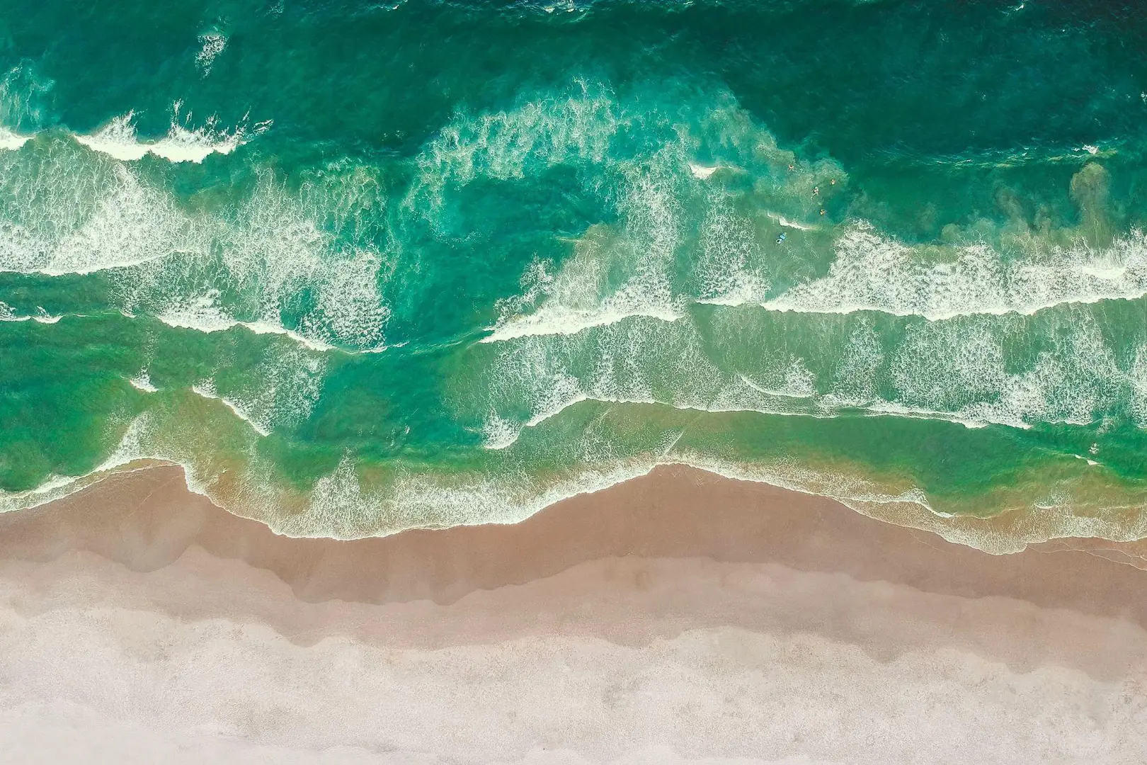 Aerial view of turquoise waves crashing onto a sandy beach with white foam, creating a natural gradient from blue to brown hues.