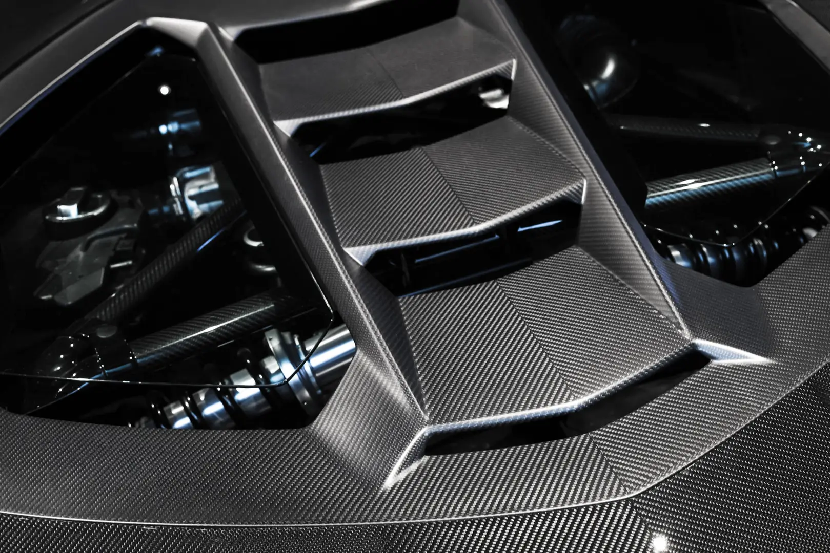 Close-up of a car's engine cover with a sleek, geometric design and visible carbon fiber texture, showcasing engine components underneath.