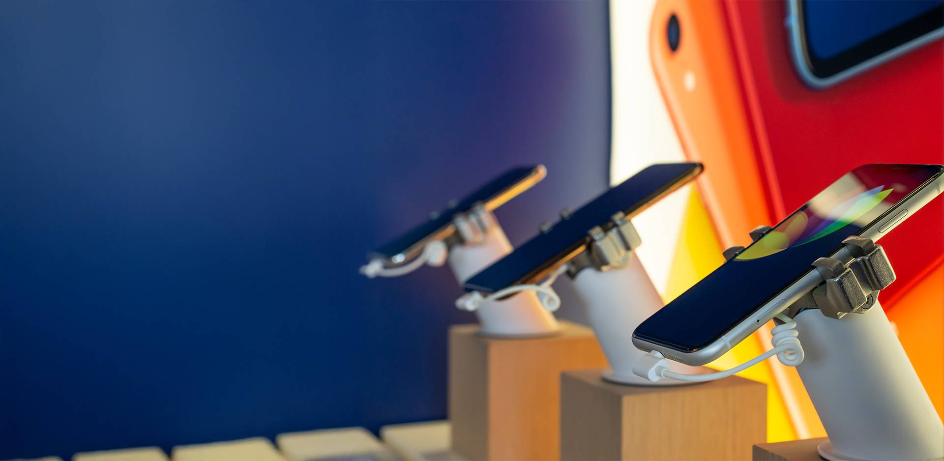 Three modern smartphones on display stands in an electronics store exhibition, in front of a colourful background.