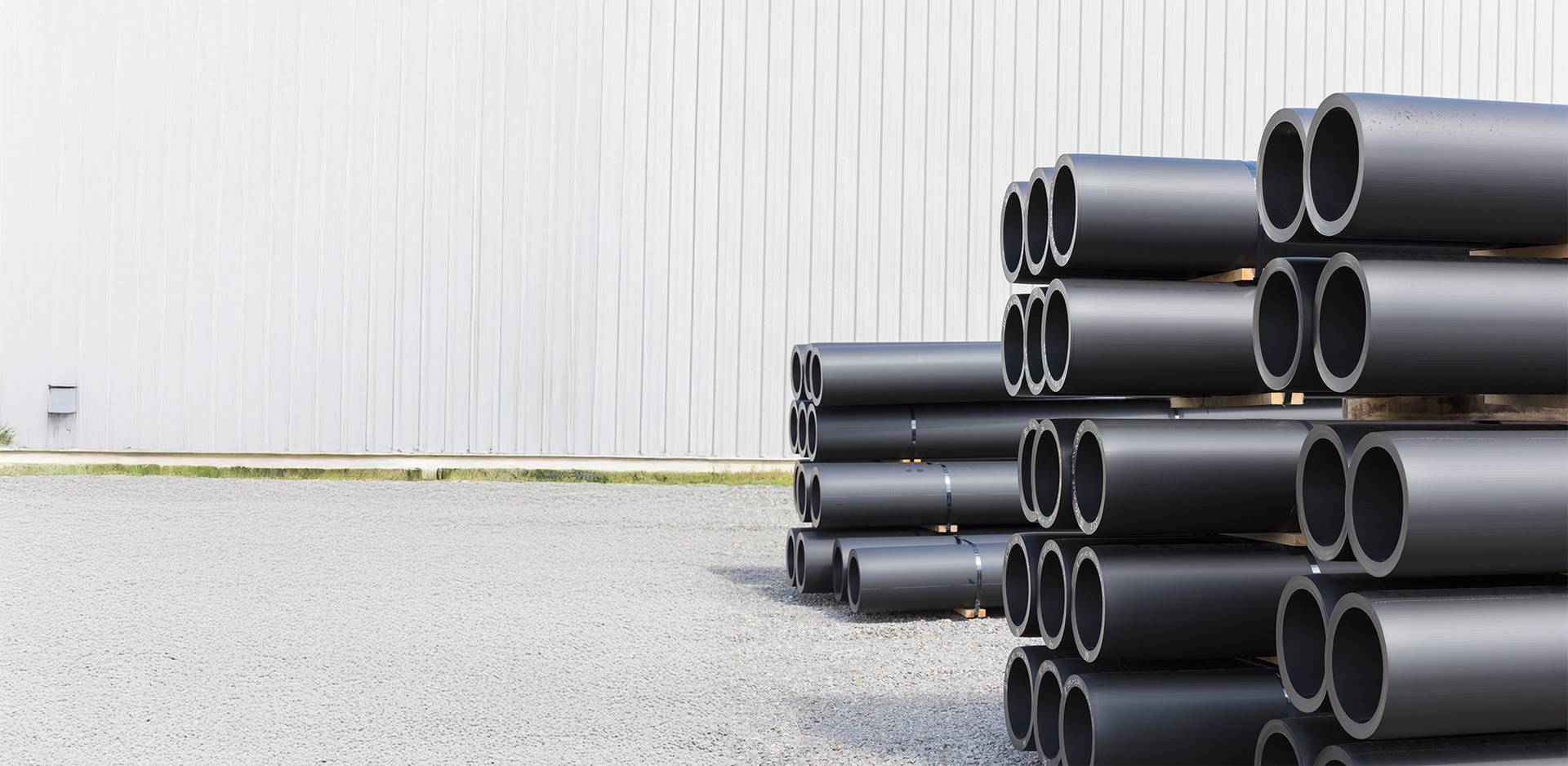 Stacked black plastic pipes next to a large warehouse on a gravel yard.