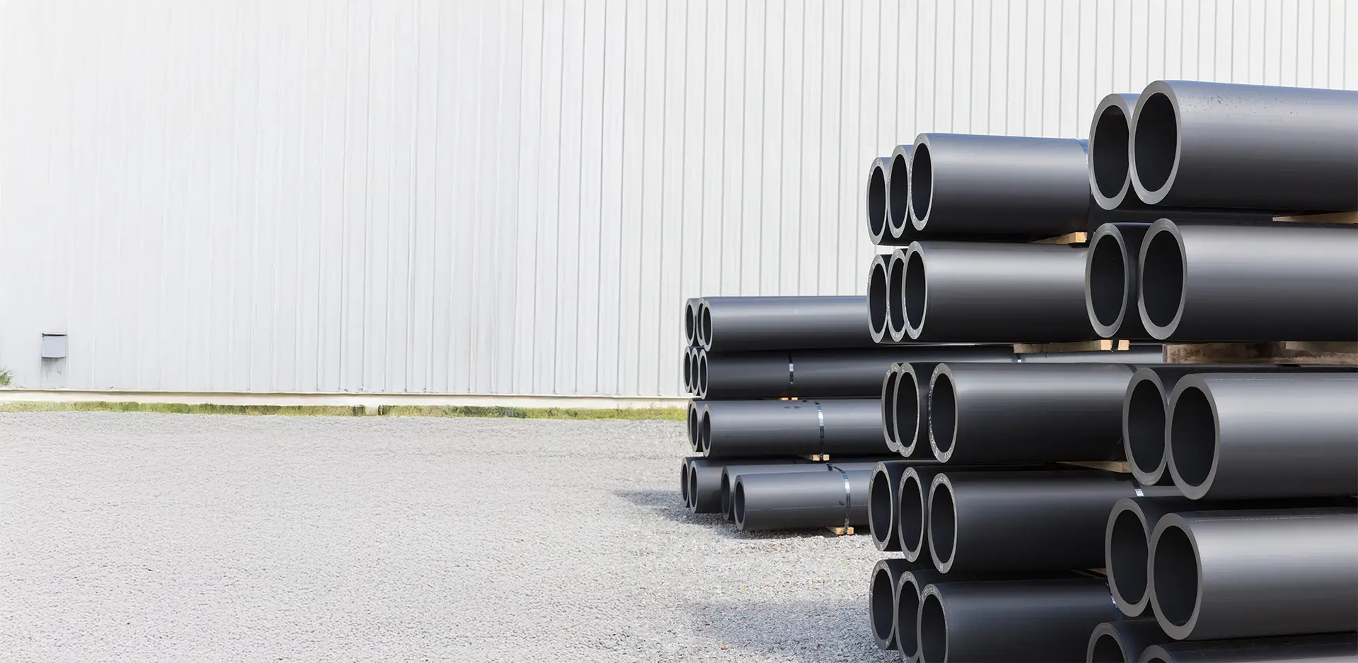 Stacked black plastic pipes next to a large warehouse on a gravel yard.