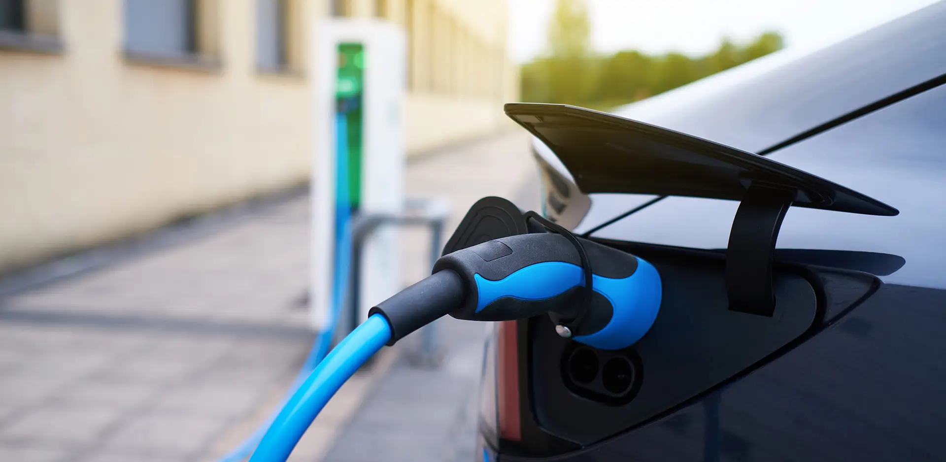 An electric car is being charged with a blue charging cable at a charging station, which is visible in the background in a car park. The image emphasises the sustainable mobility and technology of modern electric vehicles.