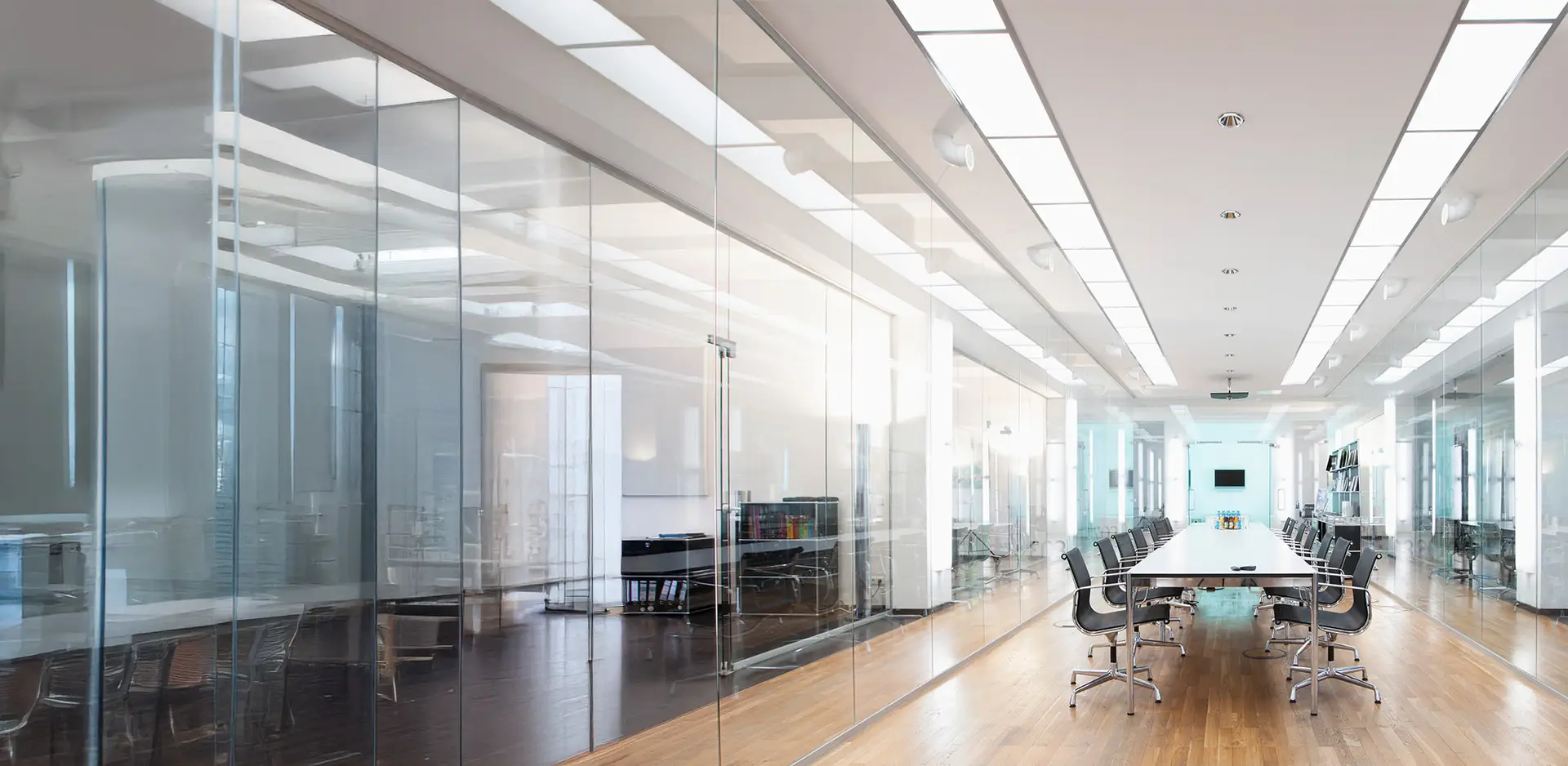 Stylish conference room with a long table and modern ceiling lighting containing PMMA material, surrounded by black chairs and enclosed by glass walls. The room is brightly lit, with contemporary office furnishings.