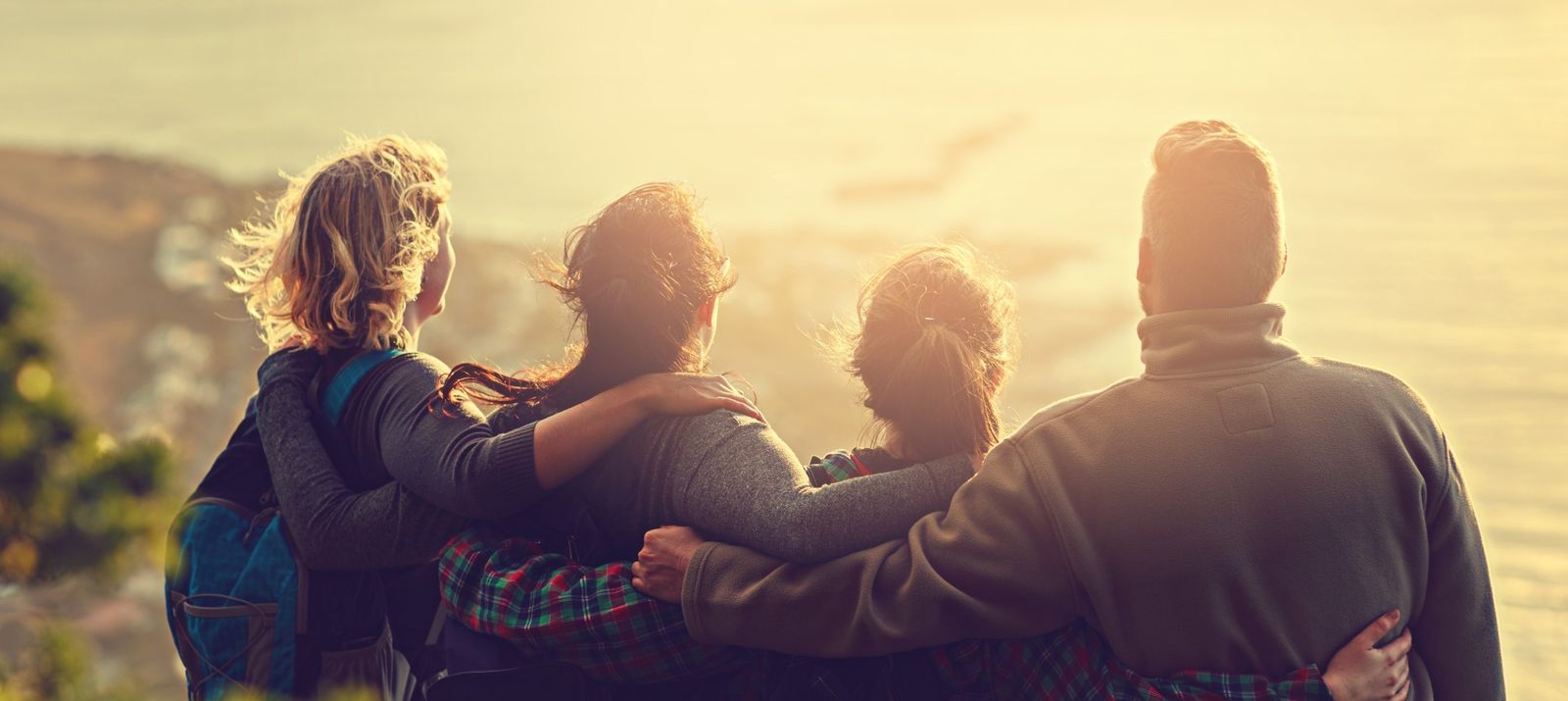 Four people with backpacks stand arm in arm, gazing at a sunlit coastal view, symbolizing friendship and adventure.