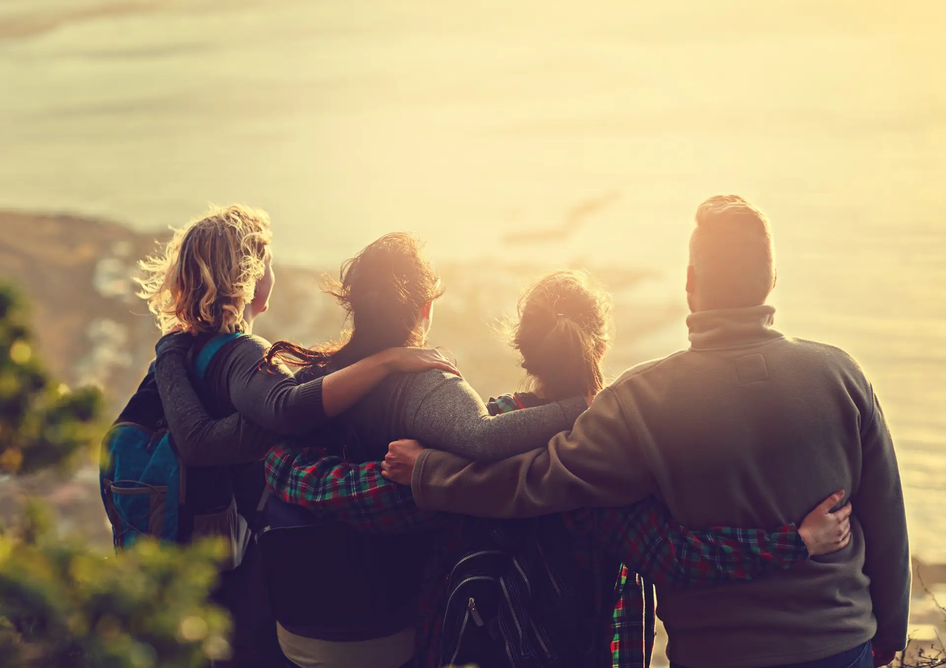 Four people with backpacks stand arm in arm, gazing at a sunlit coastal view, symbolizing friendship and adventure.