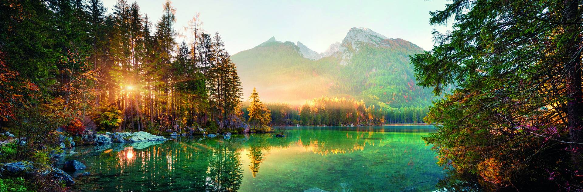 Sunrise over a lake surrounded by dense forest and mountains, with sunlight filtering through the trees and reflecting on the water.
