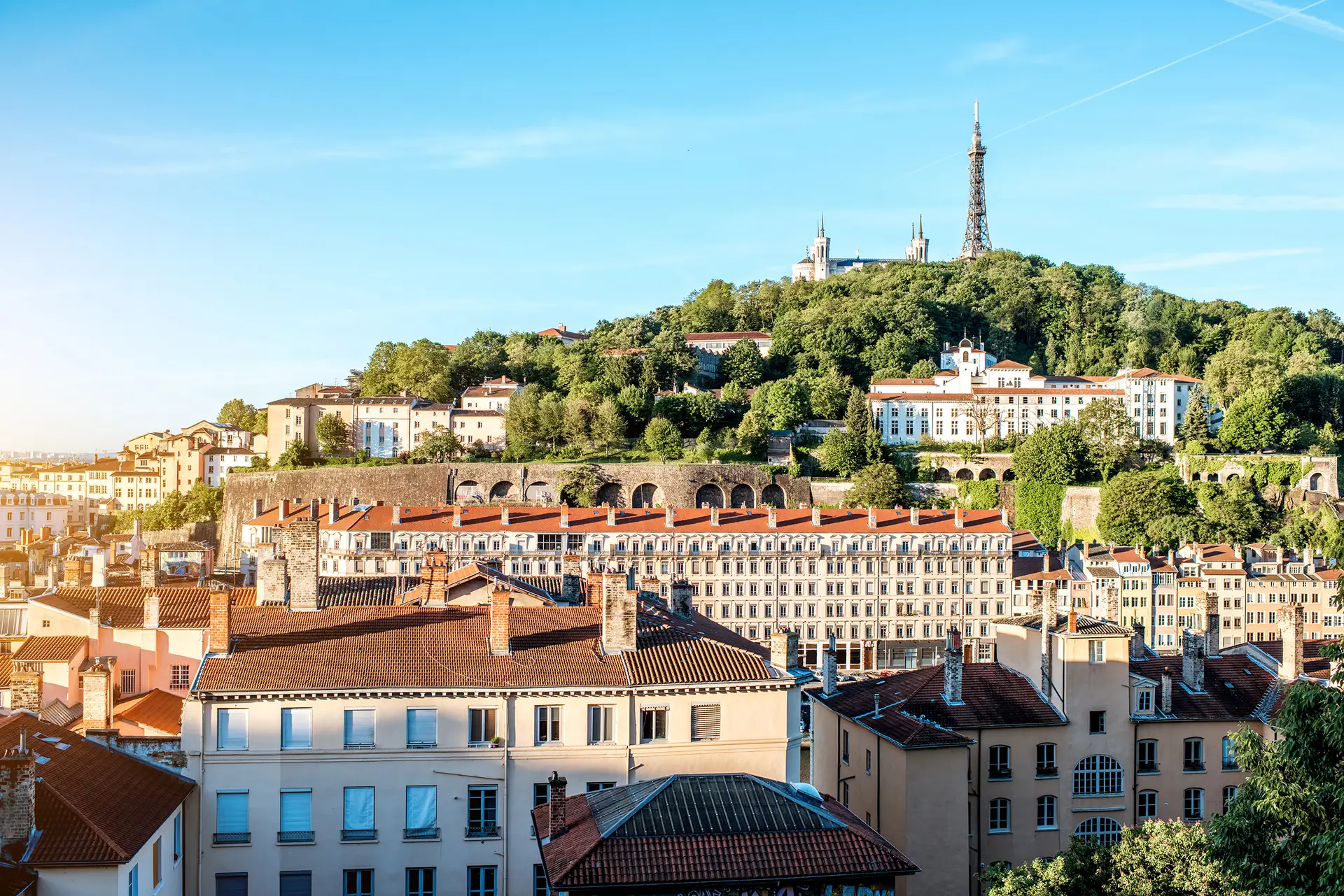Scenic view over Lyon, France, with historic buildings, lush greenery, and the Basilica of Notre-Dame de Fourvière atop the hill.