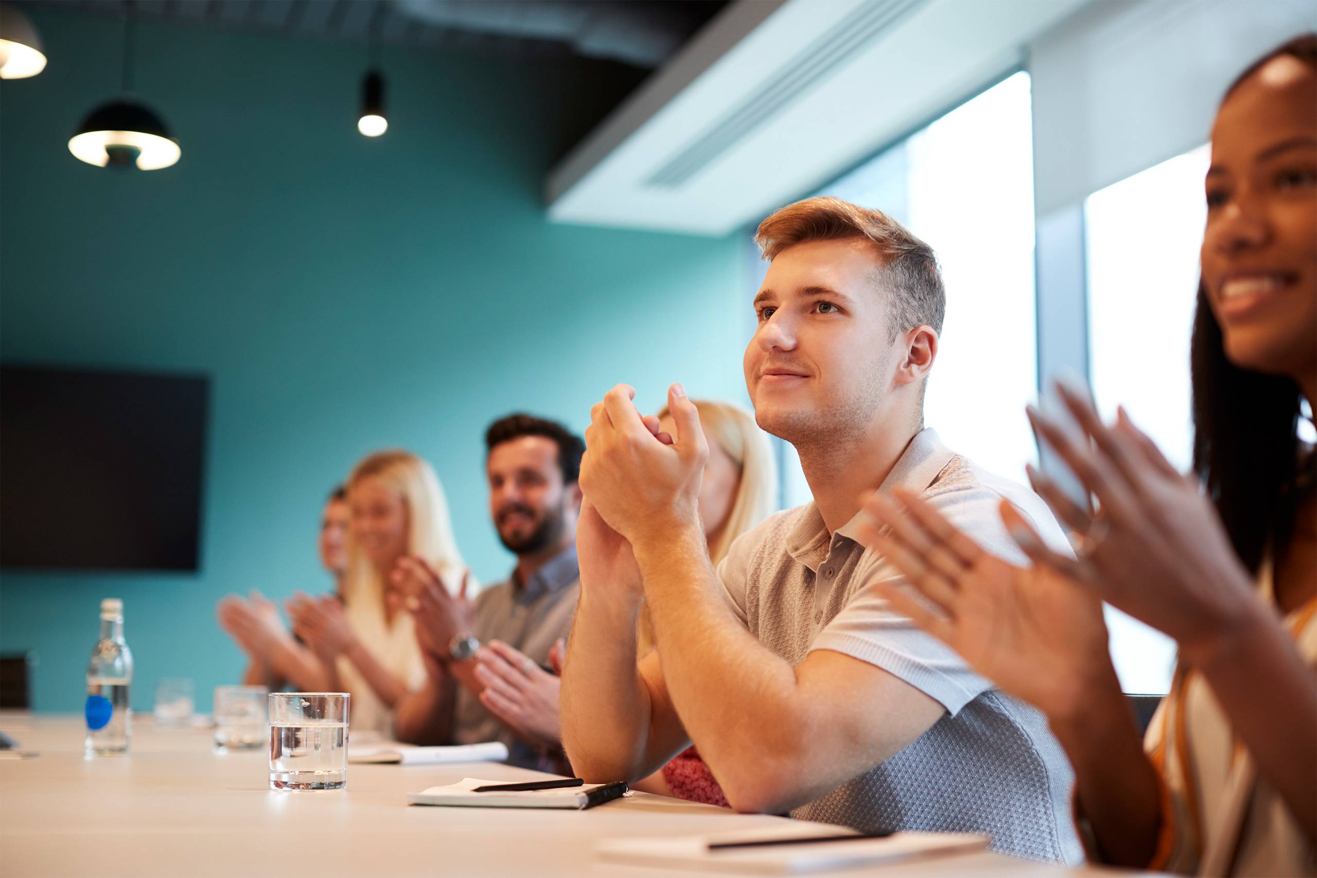 Group of young candidates sitting at the boardroom table applauding presentation at Business Graduate Recruitment Assessment Day