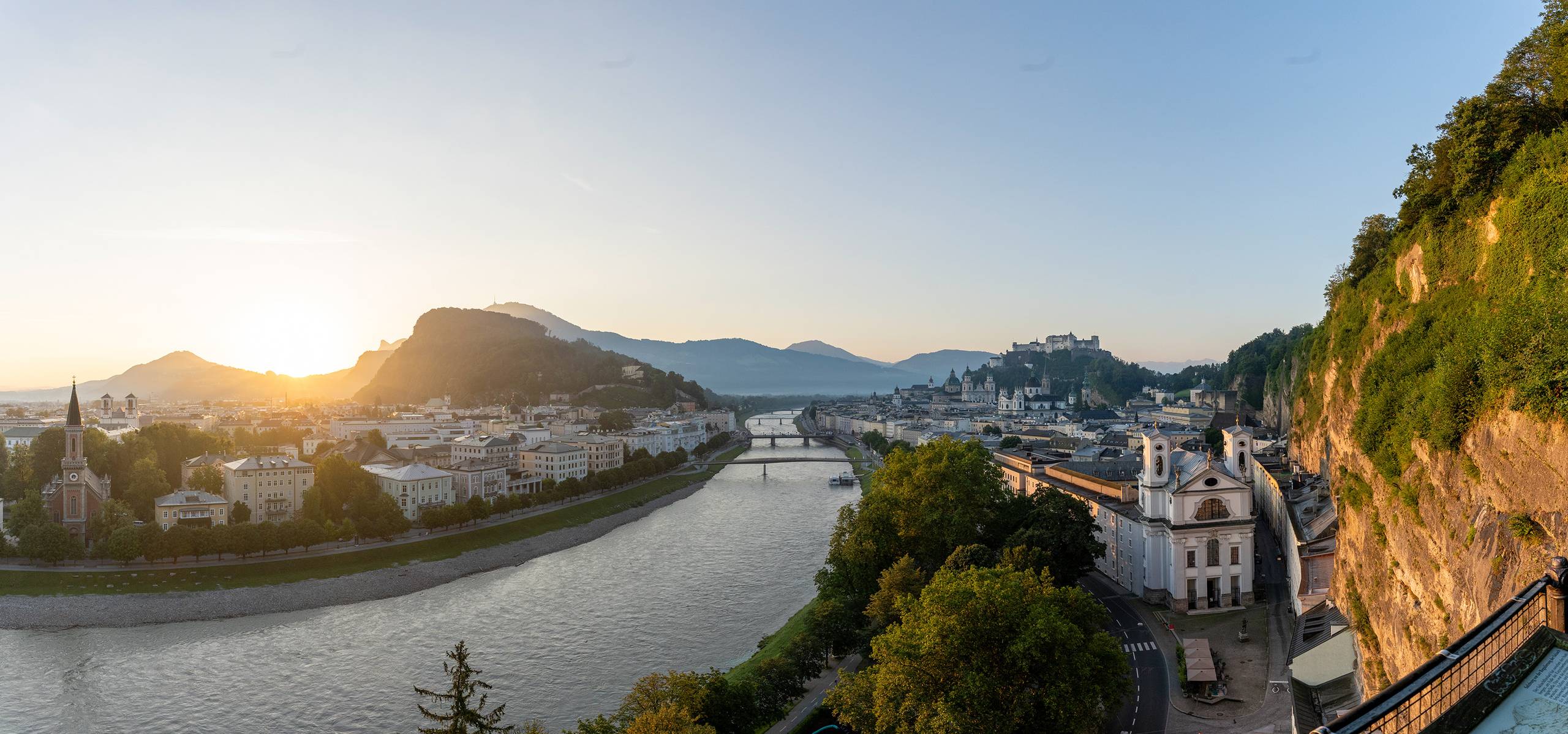 Panoramic view of Salzburg at sunset with the Salzach River, a bridge, historical buildings, and mountains in the background.
