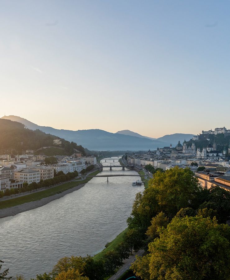 Panoramic view of Salzburg at sunset with the Salzach River, a bridge, historical buildings, and mountains in the background.