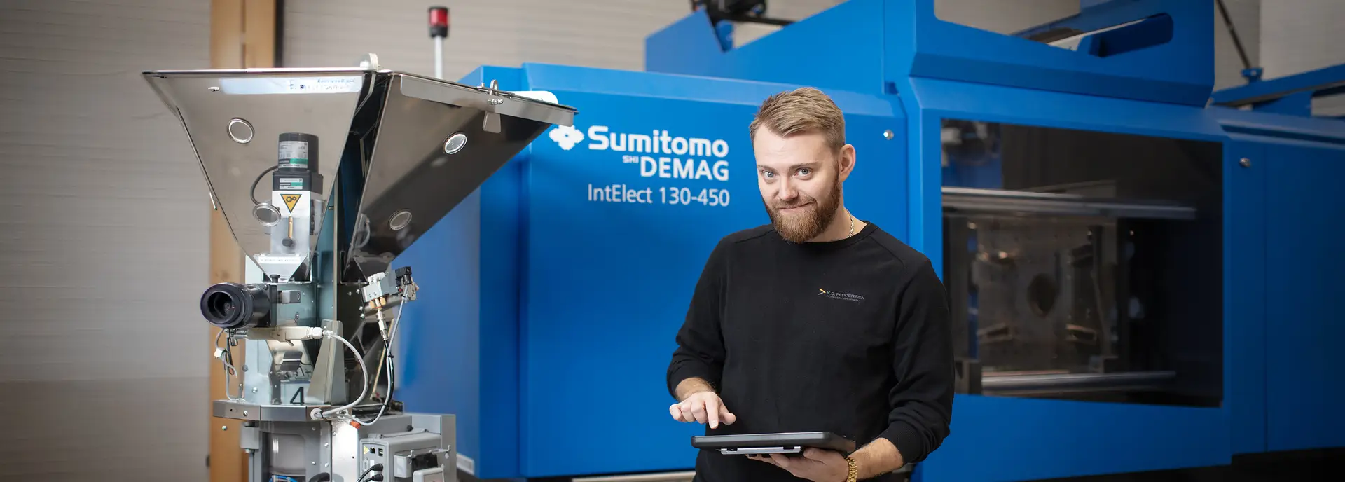 A person with a tablet stands beside industrial equipment and a blue machinery labeled "Sumitomo (SHI) Demag Injection Molding Machine."