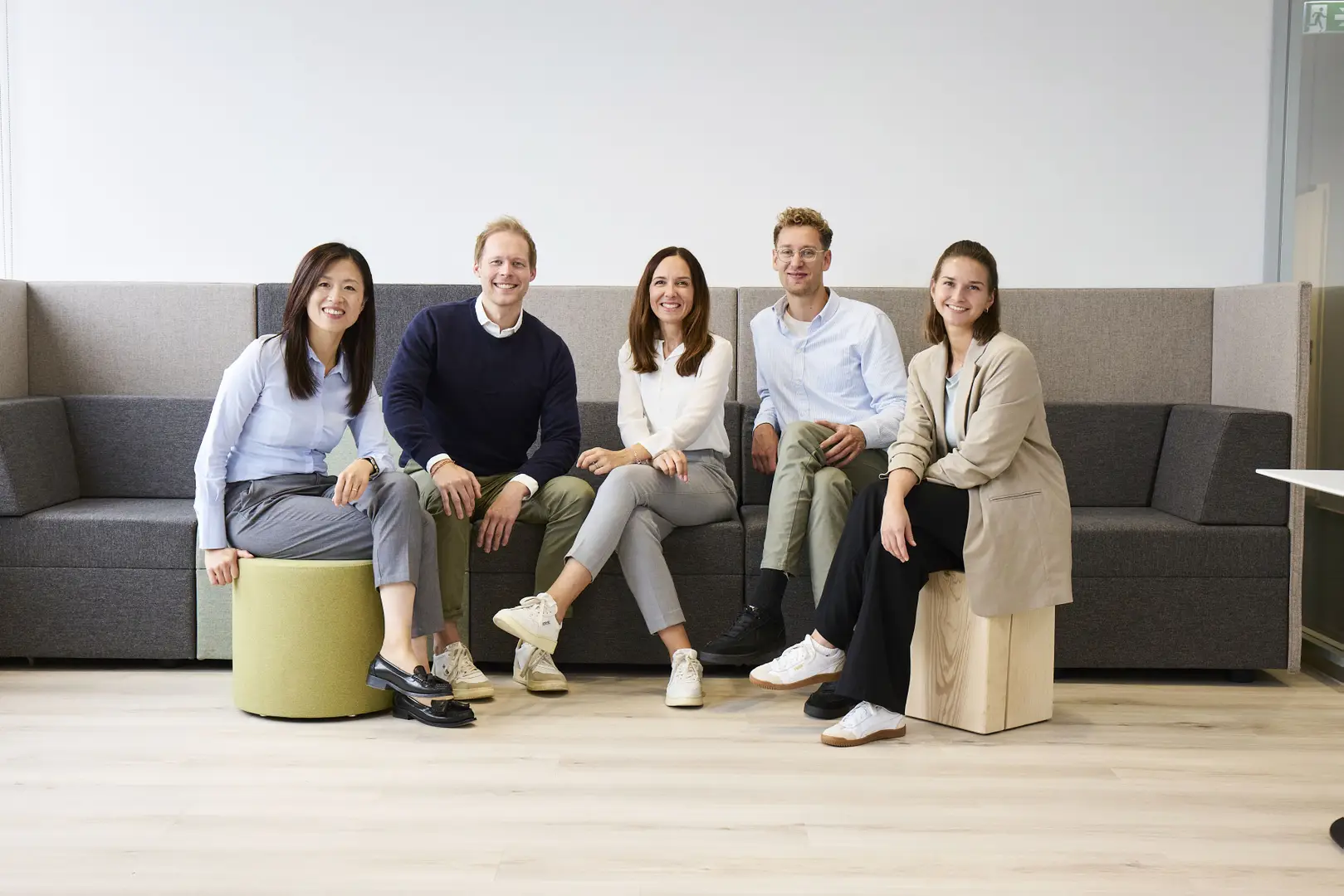 Five people sitting on stools and a couch in a bright room, smiling at the camera. They are dressed for office work.
