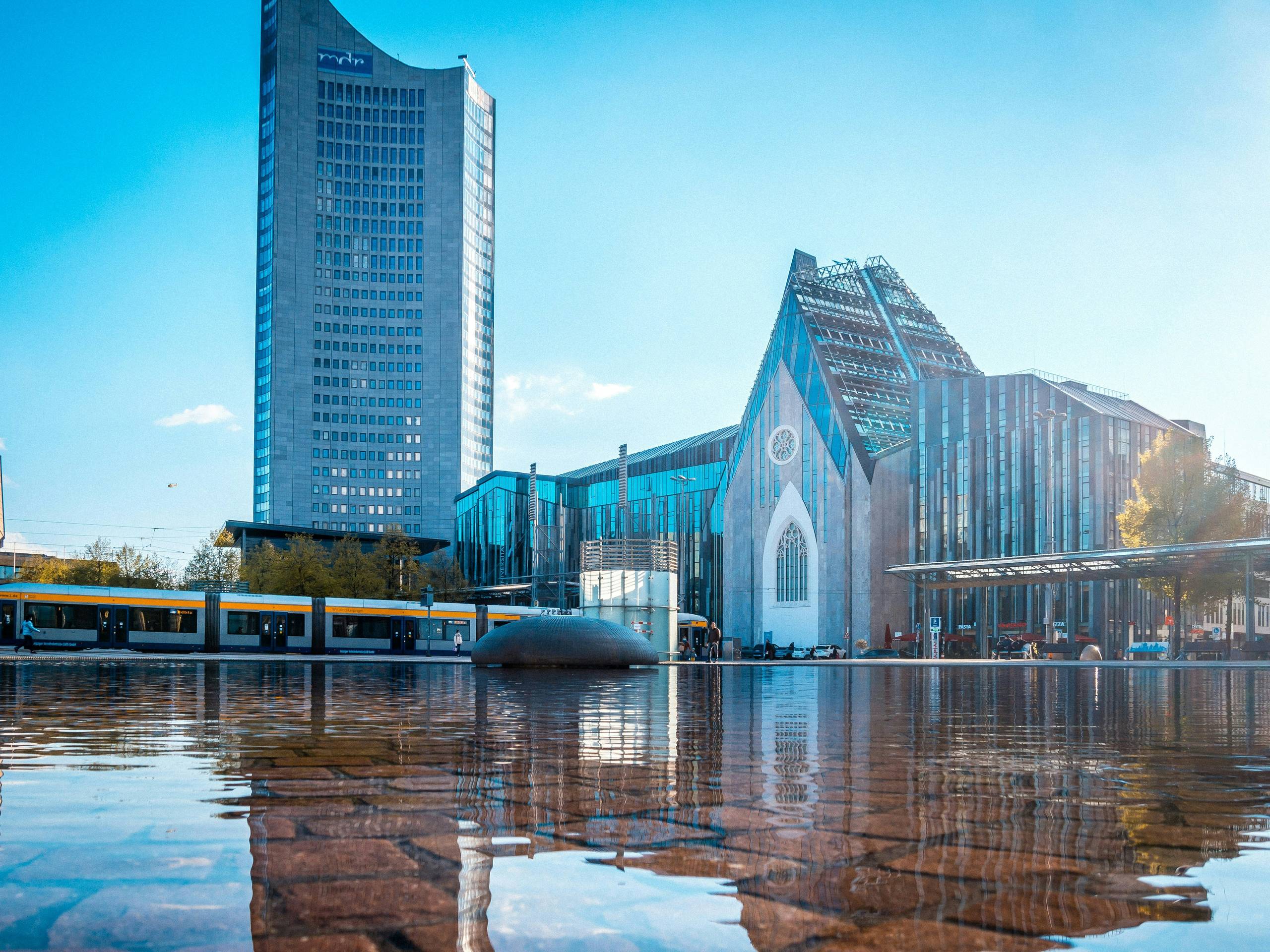 Modern buildings reflecting in a water surface under a clear blue sky. A tram and people are visible in front of the structures.