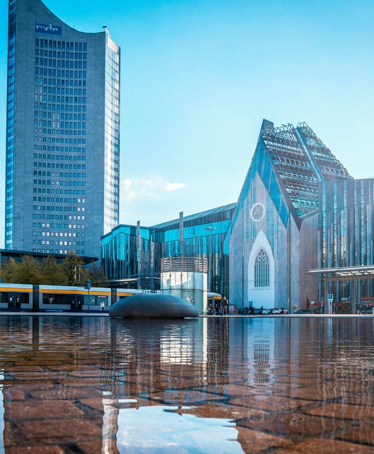 Modern buildings reflecting in a water surface under a clear blue sky. A tram and people are visible in front of the structures.