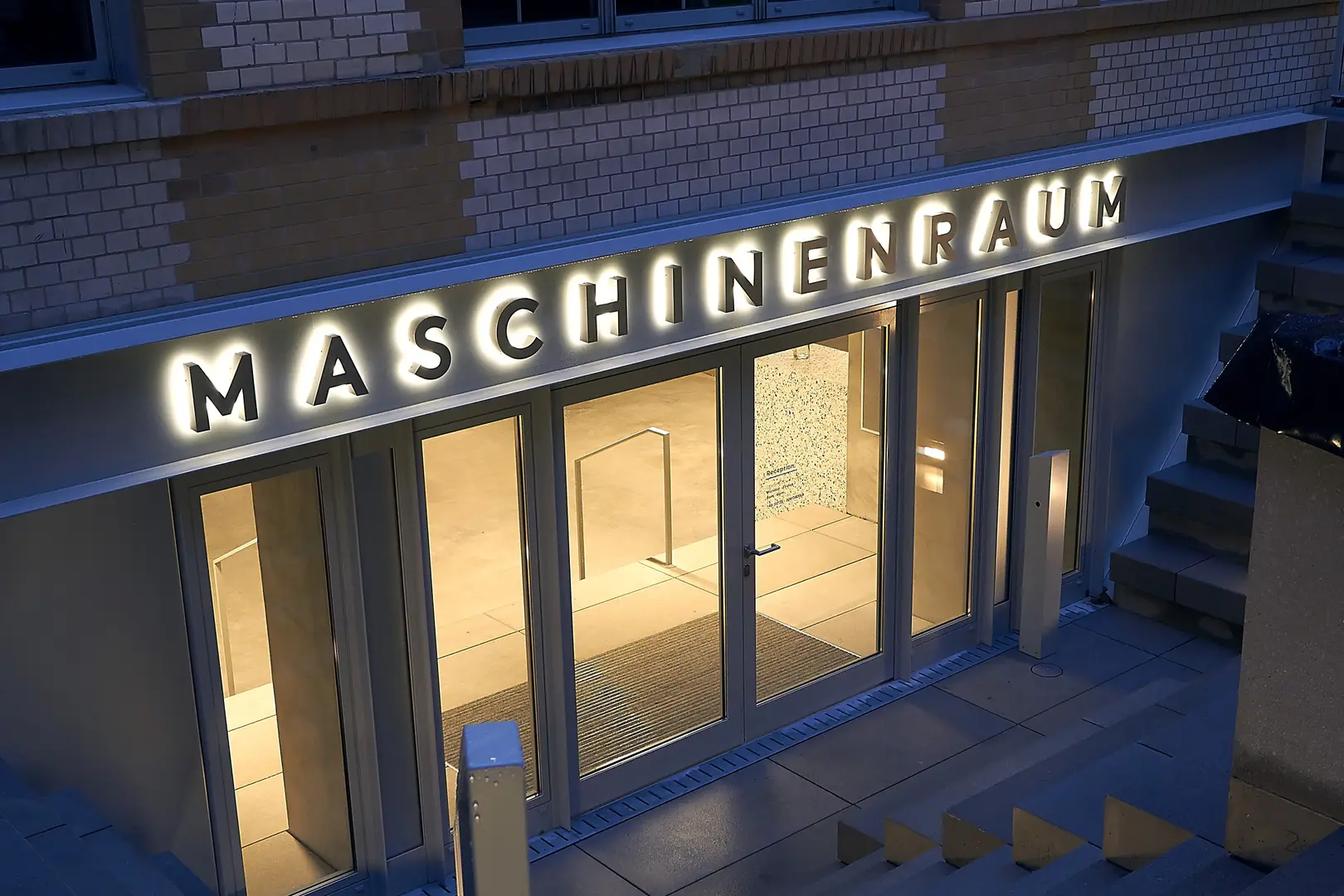 Illuminated storefront with large, glowing "Maschinenraum" sign above glass doors, set against a brick wall.