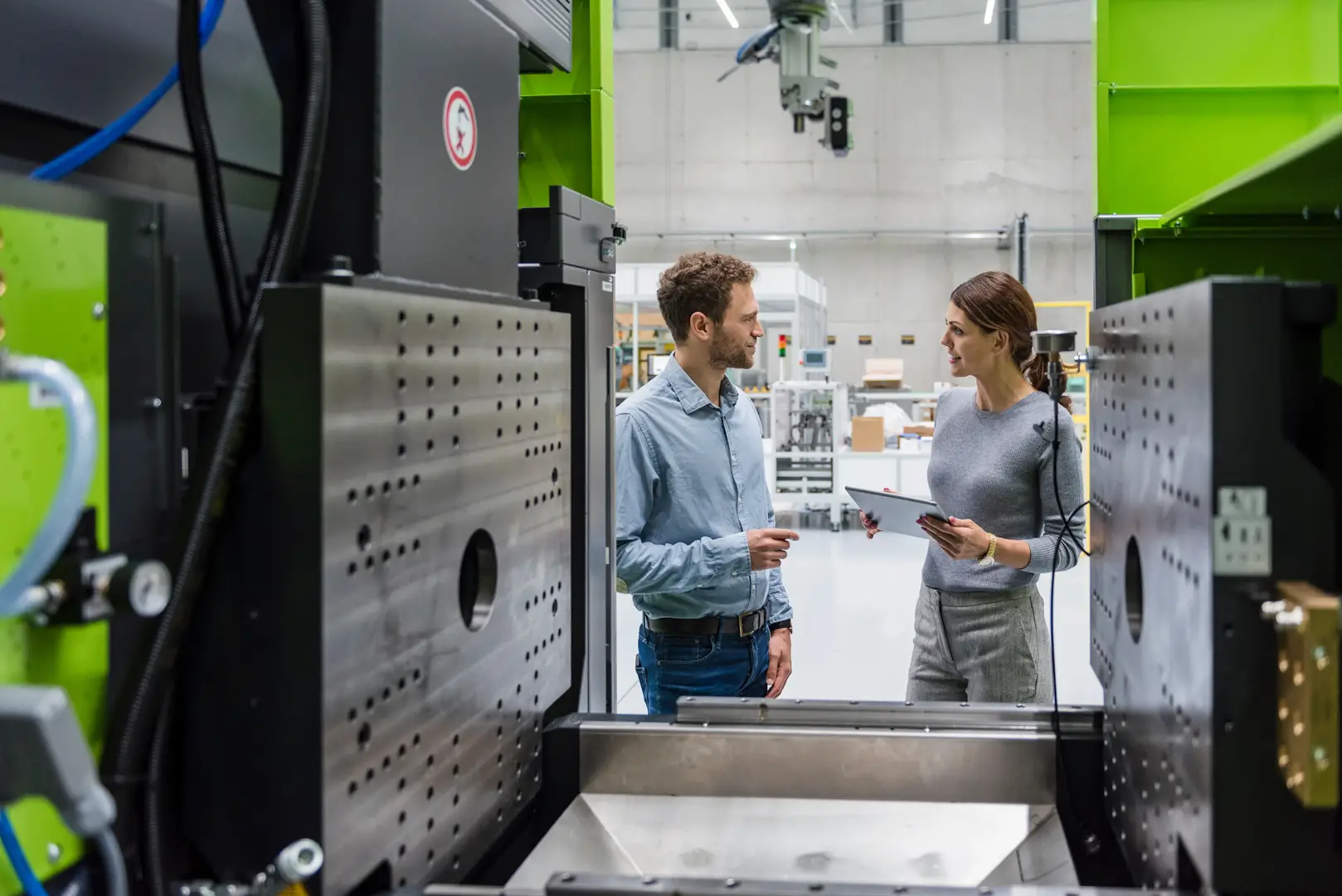 Two people in a modern industrial setting, discussing near large machinery, holding a clipboard and paper.