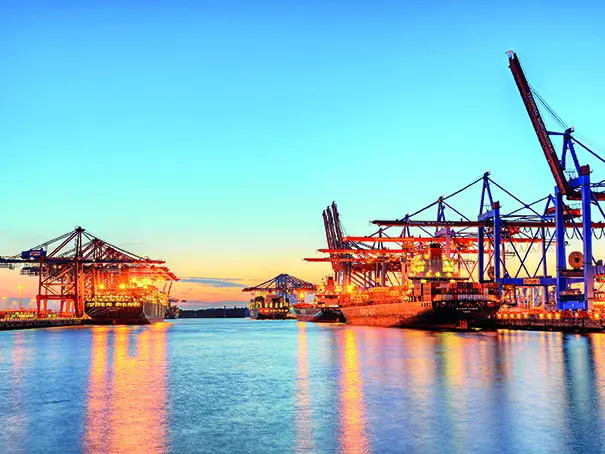 A vibrant port at sunset with illuminated cranes and ships reflecting on the water, under a clear blue sky.