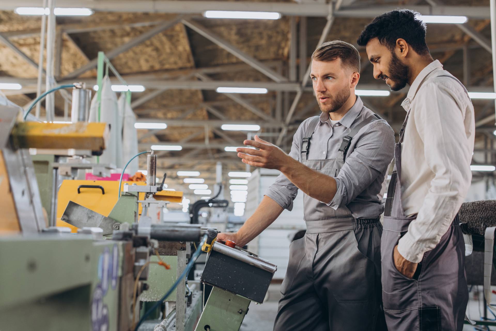 Two plastic factory workers in uniforms discuss machinery operations in an industrial setting, surrounded by equipment and tools.