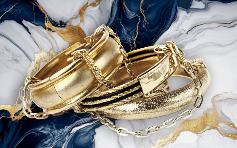 Mehrere goldene Armreifen und Ketten liegen auf einer eleganten, marmorierten Oberfläche mit blauen und goldenen Akzenten.