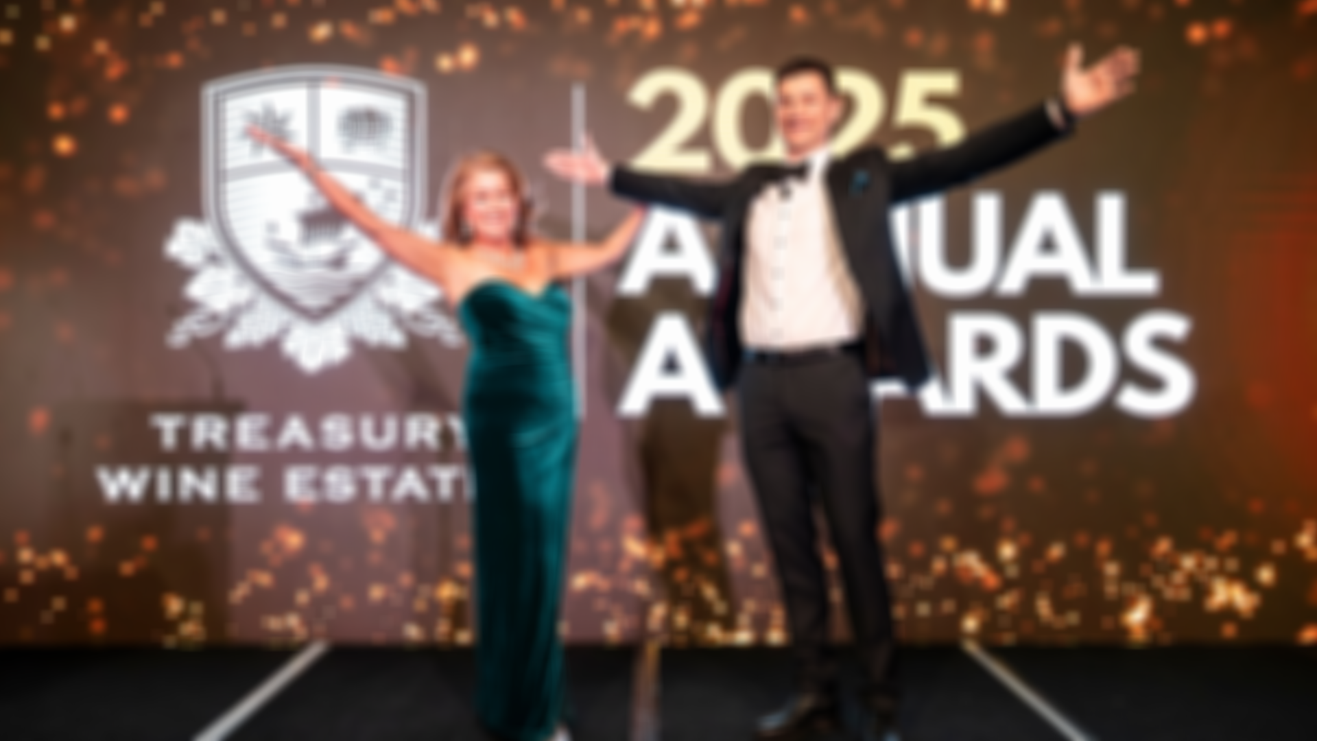 A man in a black tuxedo and a woman in a green velvet gown stand with arms outstretched on stage in front of the Treasury Wine Estates crest and the words “2025 Annual Awards.”
