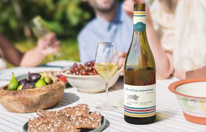 A bottle of Squealing Pig mid strength wine next to a glass and some food with people in the background enjoying a drink