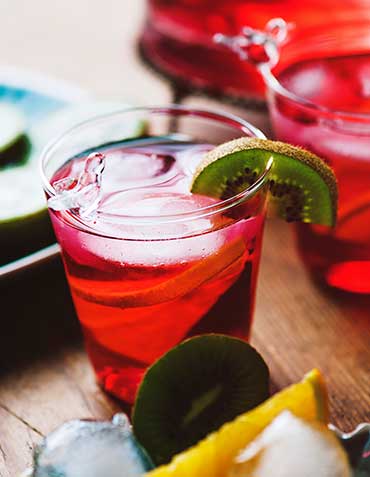 Red sangritua cocktail in short glass with a kiwi-fuit garnish on a wooden table surrounded by fruit