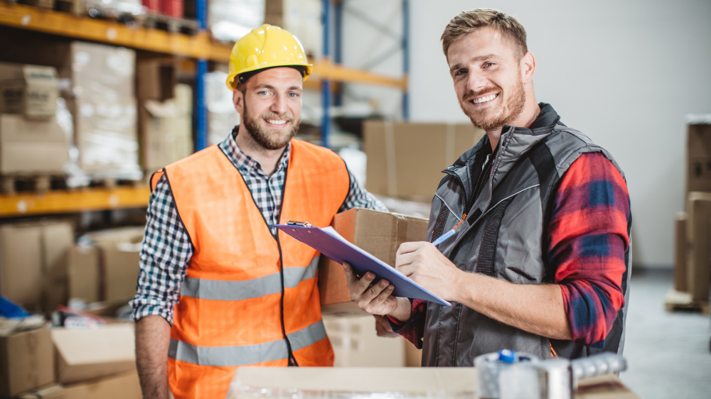 Deux employés d’entrepôt souriants dont l’un porte un gilet de sécurité et un casque avec un clipboard et l’autre une chemise à carreaux tenant un colis RTTV TMS visibilité transport en temps réel