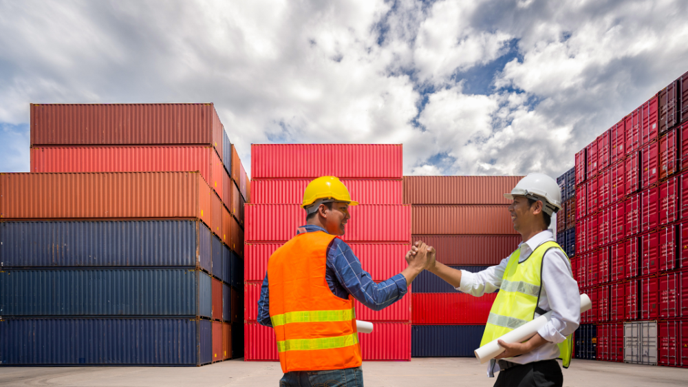 Deux travailleurs en tenue de sécurité se serrent la main devant des conteneurs empilés sous un ciel nuageux RTTV TMS visibilité transport en temps réel