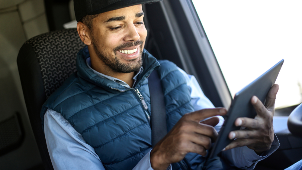 Hombre con chaleco y gorra azules usando una tablet dentro de un vehículo, consultando los tiempos de conducción y sonriendo mientras interactúa con la pantalla.