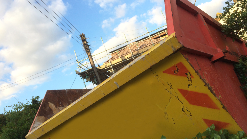 Vue en contre-plongée d’une benne jaune et rouge devant un échafaudage et un ciel bleu, illustrant le suivi bennes chantier.