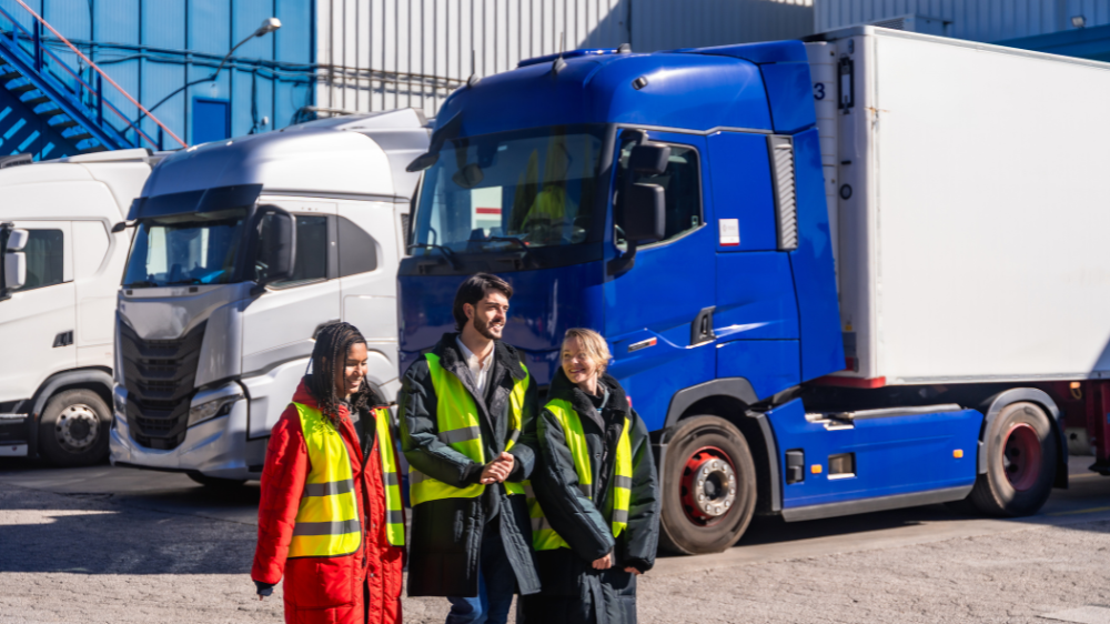 Tres personas con chalecos de alta visibilidad posan sonrientes frente a camiones articulados estacionados en una zona industrial soleada, transporte internacional de mercancías.