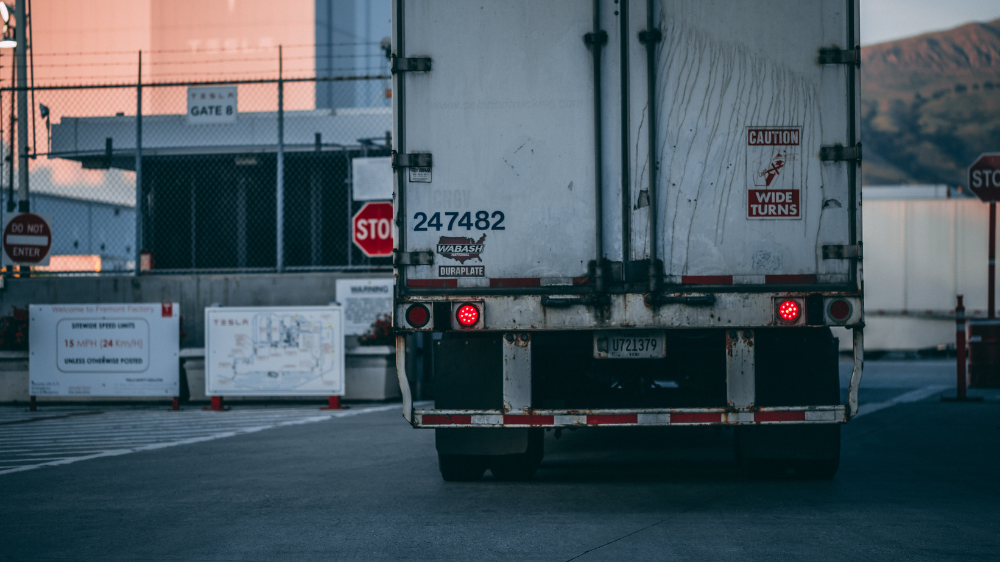Vista trasera de un camión estacionado con un cartel de “Caution: Wide Turns”, rodeado de señales de stop y edificios industriales, cuya posición puede supervisarse mediante un rastreador GPS.