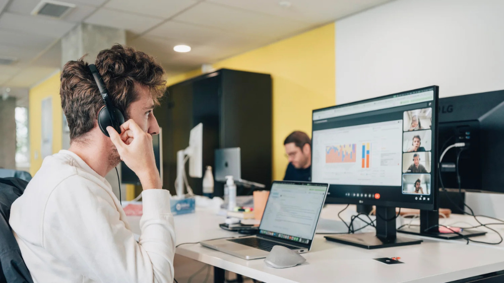Hombre con auriculares trabajando en una oficina, utilizando un software de gestión del transporte en un ordenador con videollamada y paneles de datos operativos.