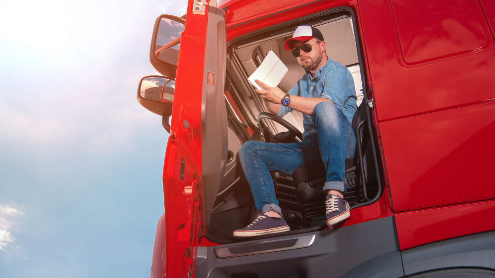 Conductor de camión con gorra y gafas de sol sentado en la cabina abierta de un tráiler rojo mientras revisa documentación, en una escena que ilustra la gestión diaria de las horas de conducción.