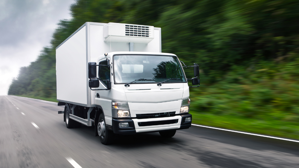 Camión frigorífico blanco circulando por una autopista rodeada de vegetación y cielo nublado, representando un transporte eficiente dentro de la logística medioambiental.