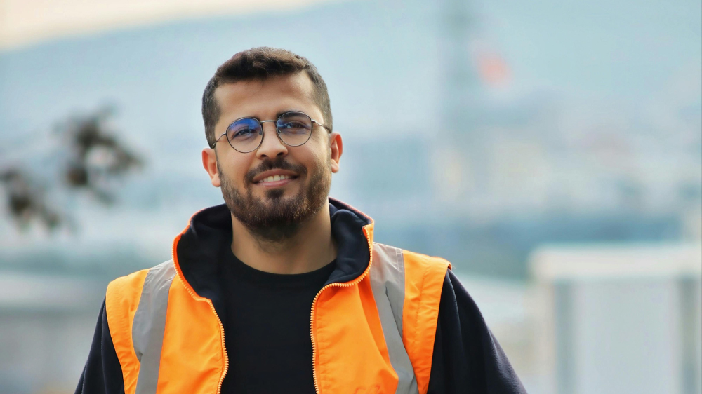 Homme souriant en gilet de sécurité orange et lunettes, debout en extérieur devant un arrière-plan industriel flou, illustrant le suivi bennes chantier sur le terrain.