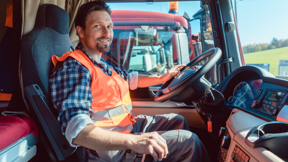 Chauffeur routier portant un gilet de sécurité orange souriant dans la cabine de son camion, avec vue extérieure lors d’une tournée suivie via un TMS transport.