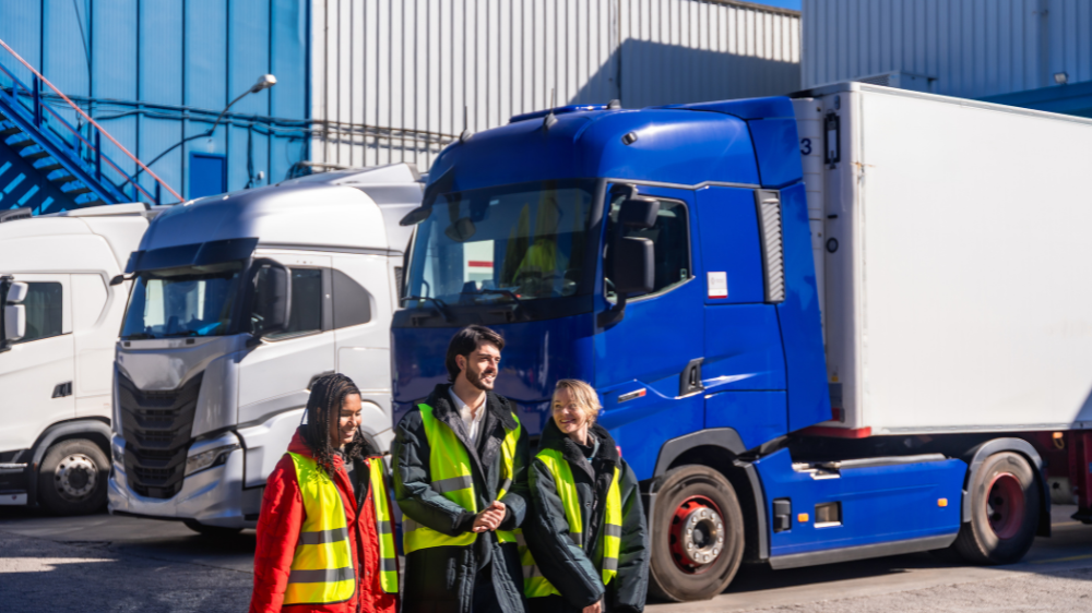 Equipo de tres operarios con chalecos reflectantes frente a camiones en un patio de carga, representando procesos organizados con programas de gestión de transporte.