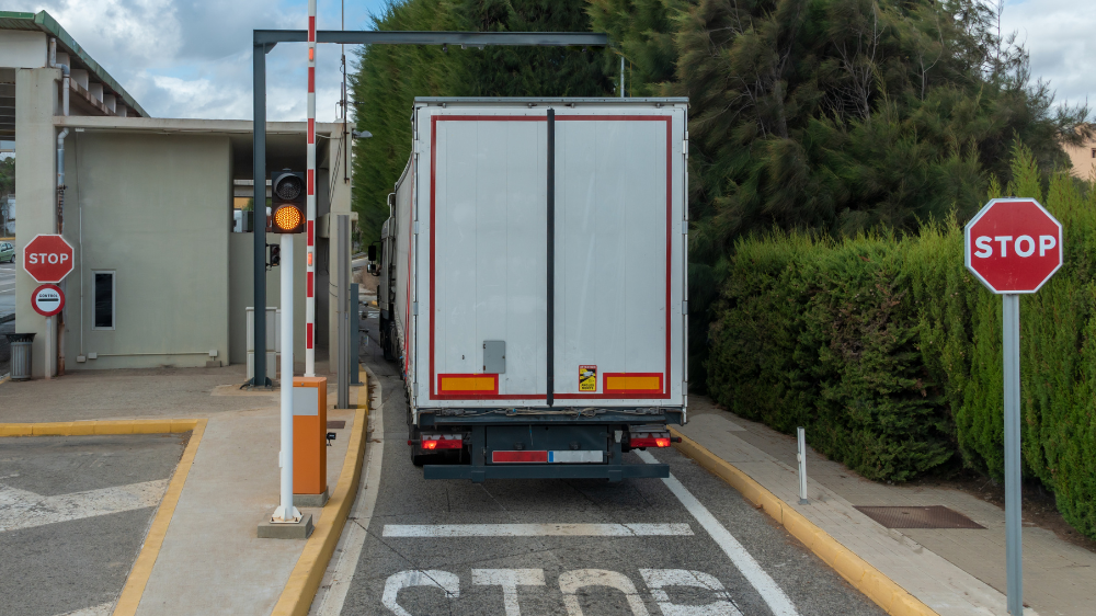 Un camión se acerca a una barrera en una entrada con control de acceso, flanqueado por señales de stop y rodeado de vegetación en un día despejado, transporte internacional de mercancías.