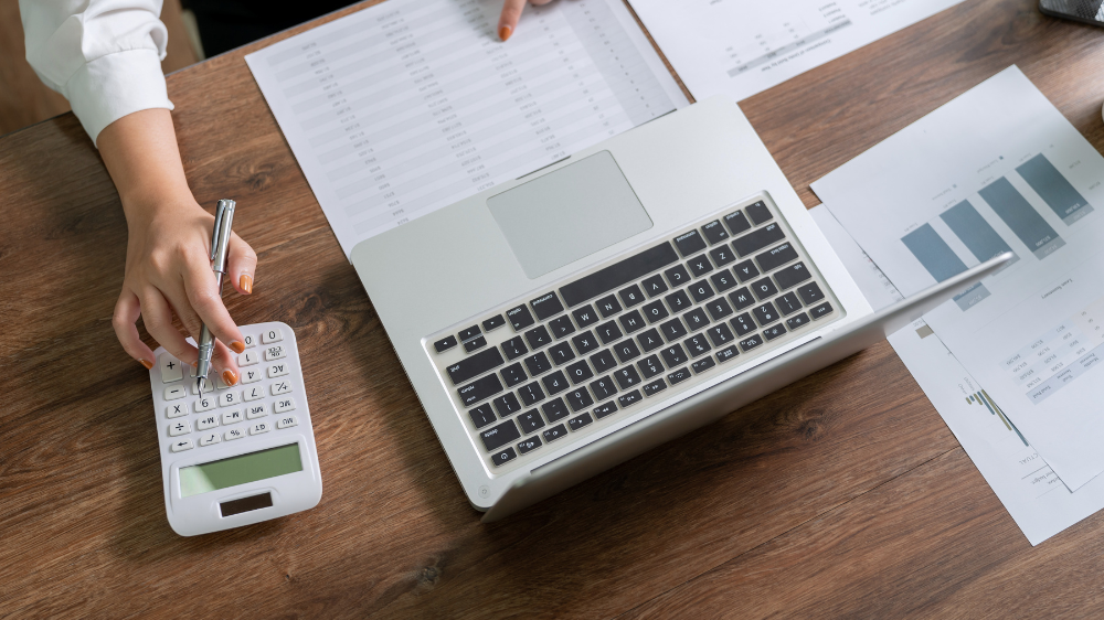 Des mains utilisent une calculatrice à côté d’un ordinateur portable et de graphiques financiers imprimés sur un bureau en bois, illustrant le calcul des coûts et le suivi des dépenses avec un logiciel d’affrètement.