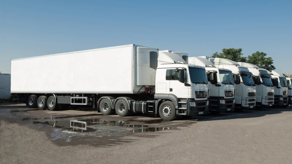 Fila de camiones blancos con remolques estacionados en un aparcamiento asfaltado bajo un cielo azul despejado, cuya ubicación puede gestionarse mediante un rastreador GPS.