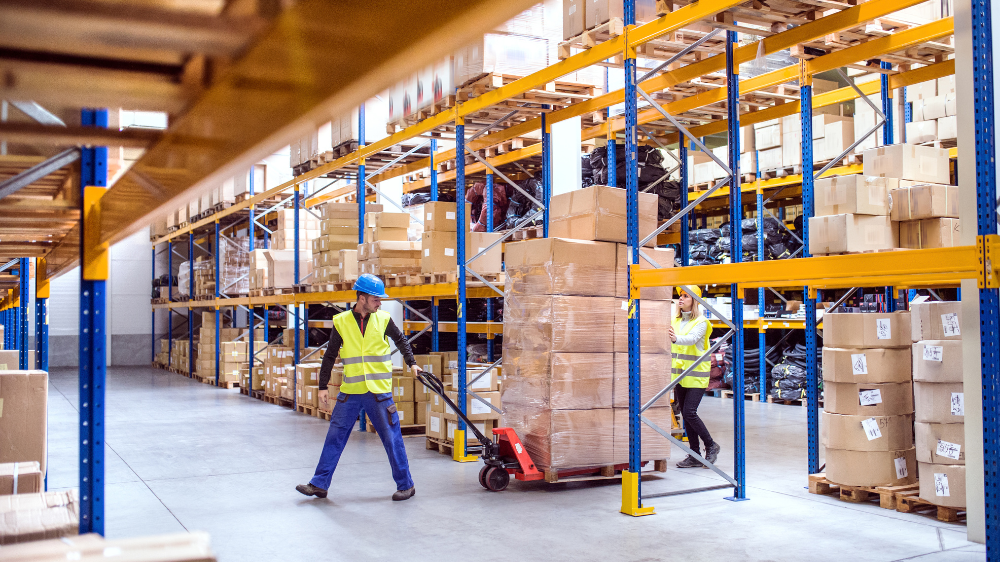 Travailleurs en gilets et casques de sécurité déplacent des cartons sur palettes avec des transpalettes dans un entrepôt 3PL logistique, entourés d’étagères remplies de colis.