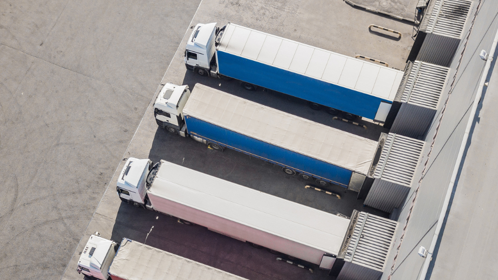 Vista aérea de cuatro camiones estacionados en un muelle de carga, listos para la estiba de carga, con remolques azules y blancos sobre una superficie de hormigón.