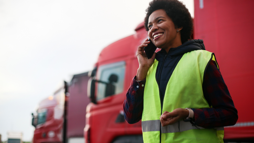 Persona con chaleco de seguridad amarillo hablando por teléfono frente a camiones rojos, coordinando una operación de estiba de carga.
