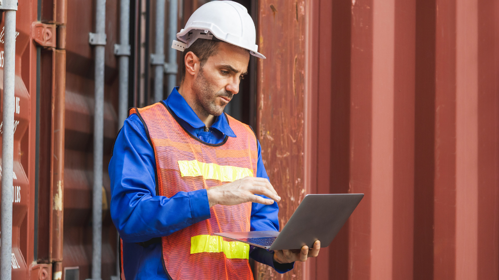 Ouvrier en casque et gilet orange réfléchissant utilisant un ordinateur portable à côté de conteneurs empilés