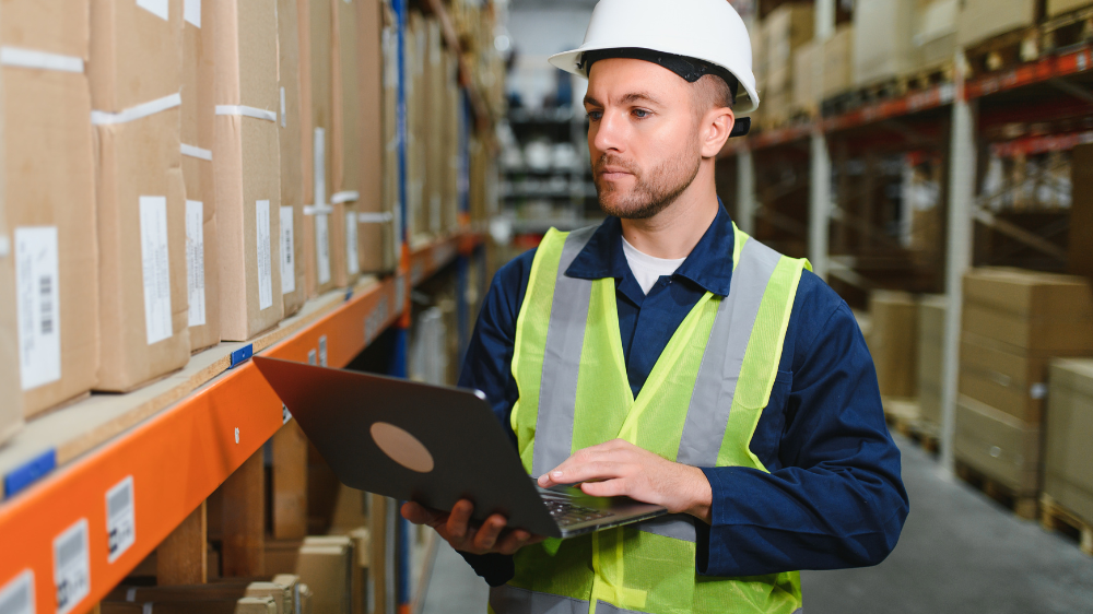 Un employé d’entrepôt portant un casque et un gilet de sécurité utilise un ordinateur portable pour vérifier les stocks sur des étagères remplies de cartons, illustrant la traçabilité et la gestion optimisée des flux via un TMS SCM.